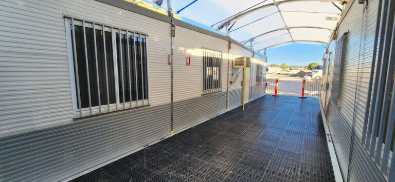 A narrow outdoor corridor between two portable buildings with a metal floor and white canopy above. The buildings have barred windows and the scene is lit by daylight. A view of a parking area and sky is visible in the background.