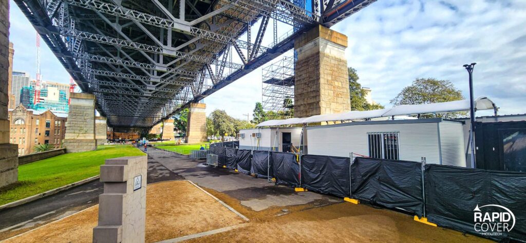 View underneath a large steel bridge with stone pillars, overlooking a construction site with temporary white buildings and black fencing. Bright green grass and trees are visible on the surrounding grounds.