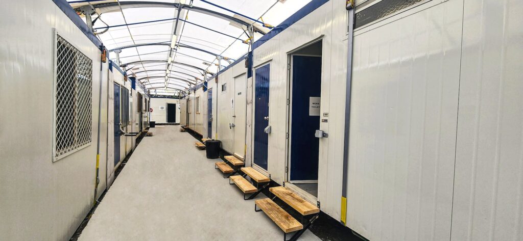 A long hallway lined with modular units on either side. Each unit has a door with a small porch and steps. The ceiling is made of a translucent material allowing natural light in. The environment is clean and organized.