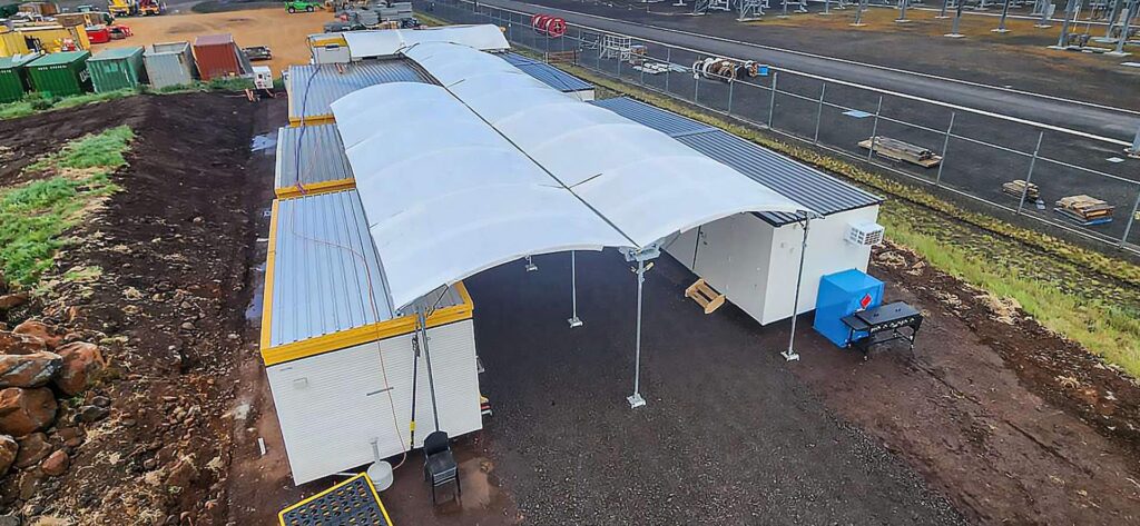 Aerial view of temporary modular structures with white canopy roofs, set on a gravel lot. Surrounding the area are various construction materials and equipment, with a fenced-off section in the background.