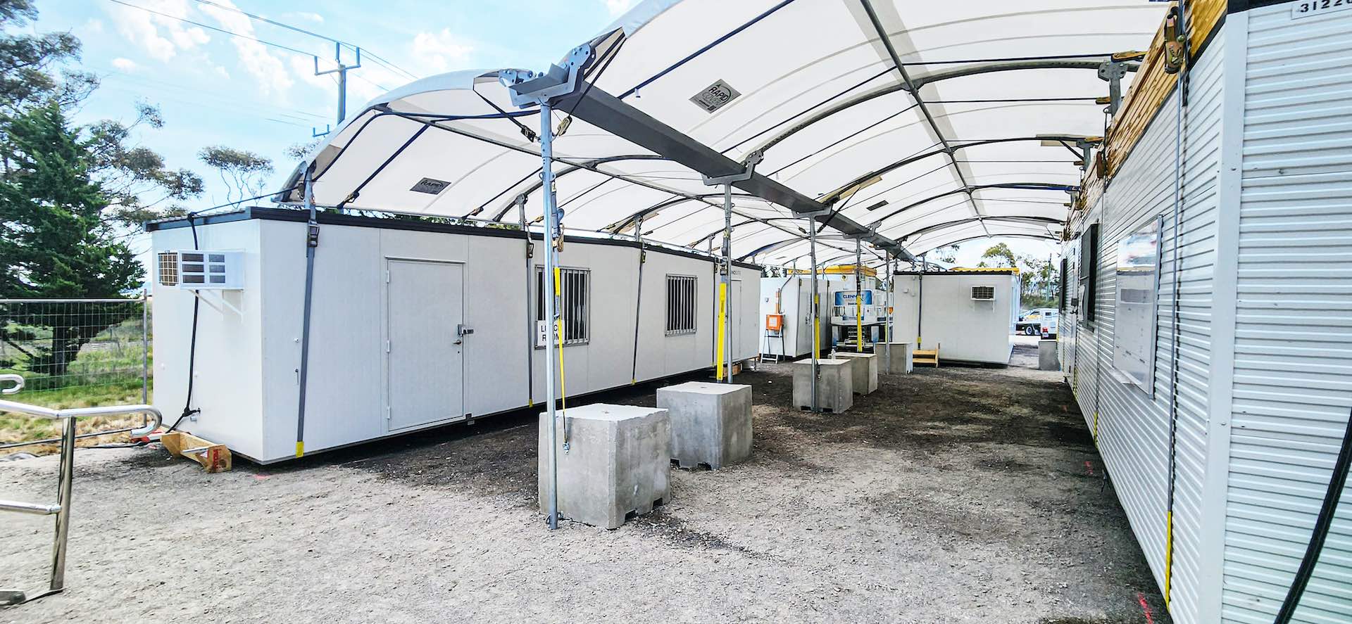 A covered outdoor area with modular buildings, possibly temporary offices or workstations. The structures are white with windows and doors. The space is shaded by an arch-shaped white canopy, and concrete blocks are placed on the ground.