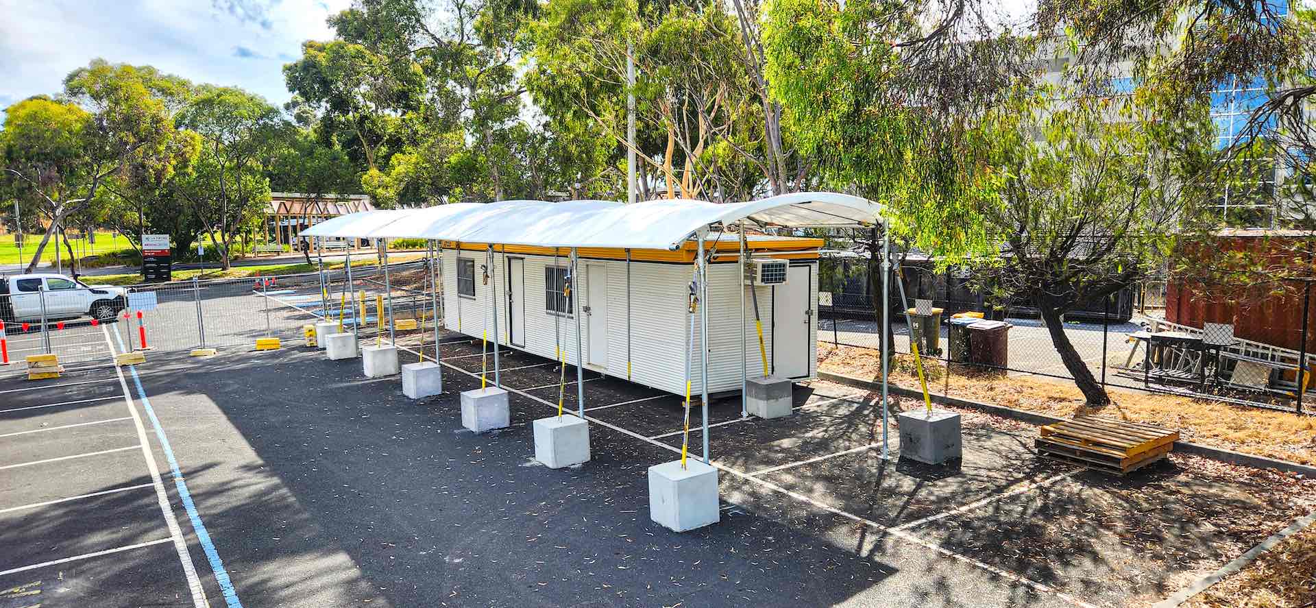 A white temporary building with a curved roof is situated in a parking lot surrounded by trees. The structure is supported by poles anchored with concrete blocks. Parking spaces are visible, along with some benches and a parked vehicle.