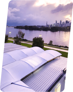 View of a river with a city skyline in the background, taken from a rooftop with curved solar panels at sunset. The sky is partly cloudy, and the water reflects the soft hues of dusk.