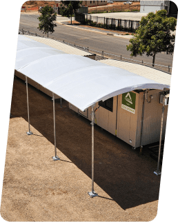 A white, curved canopy is installed over a walkway beside a long, portable building on a sunny day. The structure is supported by metal poles, with a tree and road in the background.