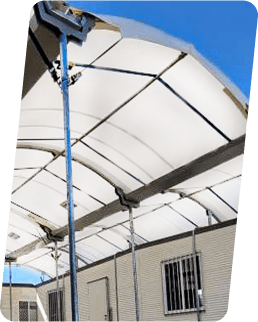 A large white canopy with metal supports is installed over a portable building against a clear blue sky. The building has small windows with bars and a door visible beneath the canopy.