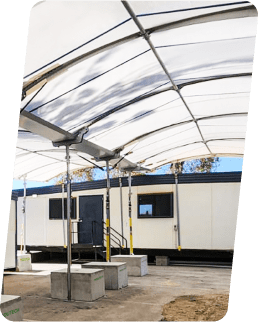 A temporary outdoor structure with a white canopy covers a walkway. In the background, there's a portable building with windows and a door. The area is supported by metal poles and surrounded by rectangular concrete blocks.